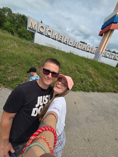 In Nikolaevka, Russia, a young couple takes a selfie in front of a large sign that reads "MOSKVA-VLADIVOSTOK". The man, wearing sunglasses and a black t-shirt with "JUST DO IT" printed on it, smiles at the camera. The woman next to him, wearing a pink cap and a white t-shirt, laughs. A young boy in a black cap and a tie-dye t-shirt is visible in the background, walking up a grassy hill. The sign is positioned on the side of a road, with a vibrant green hill and trees behind it. A colorful, abstract sculpture stands to the right of the sign, with layers of red, blue, and white. The sky is cloudy, suggesting an overcast day.