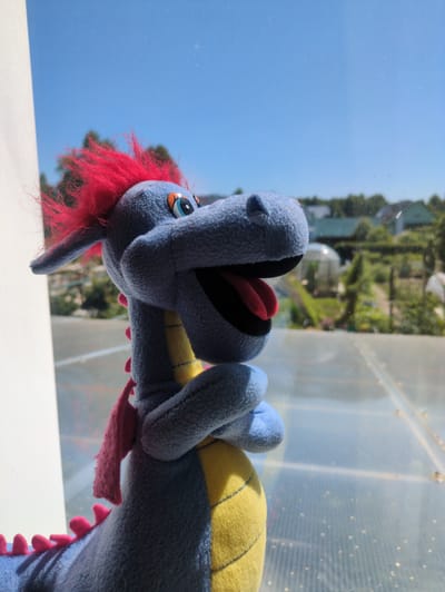 The image features a close-up, tilted shot of a blue stuffed dragon toy. The dragon has bright red, spiky hair, a yellow and blue striped back, and large blue paws. Its mouth is open, revealing a red tongue and black lining. The background is a blurred, sunlit scene with a clear blue sky, green trees, and a translucent, ribbed structure that appears to be made of plastic or glass. The lighting suggests it is daytime, possibly mid-morning or afternoon, with bright sunlight. The overall impression is a whimsical and possibly playful scene, perhaps set outdoors or near a window, given the light and background elements. There is no visible text in the image.