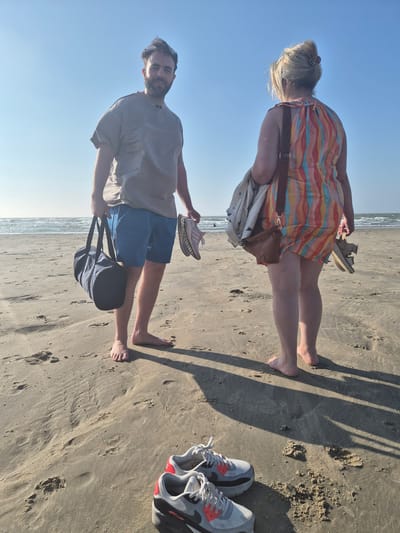 Two individuals are standing on a sandy beach with the ocean and a clear blue sky in the background. The man, on the left, is wearing a grey t-shirt and blue shorts. He is holding a dark blue duffel bag in his left hand and a pair of pink and white sneakers in his right hand. The woman, on the right, is facing away from the camera and is wearing a colorful striped dress. She is carrying a white jacket over her left arm and a brown purse slung across her body. In her right hand, she is holding a pair of beige sandals. In the foreground, a pair of grey and red Nike Air Max sneakers are placed on the sand. The image appears to be taken in the city of Blankenberge, Belgium, based on the provided context. The scene depicts a casual beach setting.