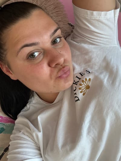 A woman is lying down, with her head tilted back and looking up at the camera. She is wearing a white t-shirt with a graphic of a daisy and the word "DICKIES" printed on it. The t-shirt is a light, casual style, and the graphic is centered on the chest area of the shirt. She has dark hair and light skin, with some freckles visible on her cheeks. Her eyes are green and her eyebrows are dark and well-defined. Her lips are full and slightly pursed. The background appears to be a bed or couch, with a pink fuzzy pillow visible to the left of her head. The overall impression is a casual, relaxed moment, possibly taken at home. The time of day and weather are not discernible from the image. There is no specific information about the location in Bulgaria, Rogosh.