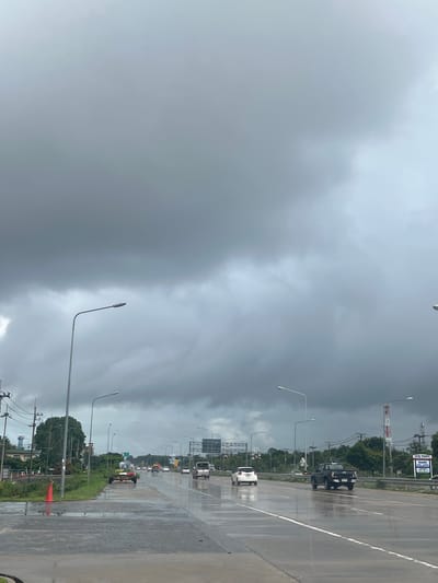 The image depicts a wet, overcast day on a highway in บ้านหนองน้ำเต้าลอย, Thailand. The sky is filled with dark, ominous clouds. The road surface is wet, reflecting the sky and vehicles. There are vehicles on the road, including a white car, a black pickup truck, and other unidentified vehicles further down the road. There are streetlights along the side of the road. Beyond the road, there is green vegetation and utility poles. There are also road signs visible.
