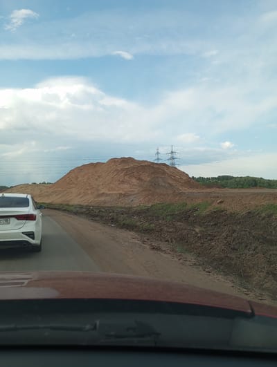 The image, taken from inside a car, shows a large mound of dirt or sand next to a road. The back of a white car is visible on the left side of the road. Power lines are present in the background, along with trees in the distance. The sky is cloudy. The vehicle's hood and windshield are visible in the foreground. The presence of Russian license plates, along with the explicit statement in the prompt, suggest the location is likely somewhere near the city of Kabanikha, Russia. Exact location is not discernible.