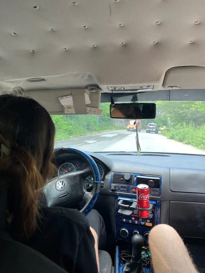 The image, taken from inside a car, shows the perspective of a passenger looking towards the driver and the road ahead. The driver, a person with long brown hair, is partially visible from the back. The car's interior is detailed, featuring a Volkswagen steering wheel with a blue patterned cover, a dashboard with air vents and a car stereo, and a can of Coca-Cola in a cup holder. A rearview mirror reflects the road, where an orange van, a dark-colored SUV, and a yellow car are visible. The exterior environment appears to be a road surrounded by lush green trees, suggesting a rural or suburban setting. The presence of the Coca-Cola can and air freshener hanging from the rearview mirror indicate the car is in use. There is no specific indication of the location being in the city of Vlahovo, Bulgaria.