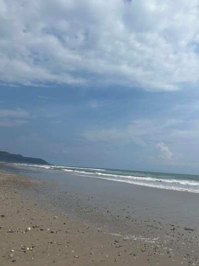 The image shows a sandy beach with a line of foamy waves in the distance, and a hill covered with vegetation in the background. The beach is covered with small rocks and seashells. The sky is bright blue with white fluffy clouds. The location is a beach in Santa Fe, Costa Rica, with coordinates 9.641 latitude, -85.168 longitude. The beach is empty with no people present.