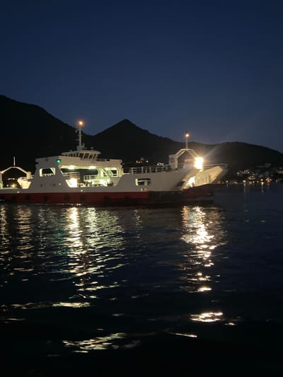 A ferry, identified by the name "GREBALI" on its side, is illuminated at night on a body of water, likely a bay or strait, near the town of Đurići, Montenegro. The ferry has a white upper structure with red trim along its waterline. Lights are visible on the ferry's decks and on a mast. The water reflects the ferry's lights, creating a shimmering effect. In the background, dark, silhouetted hills or mountains rise, with scattered lights indicating a settlement on the shore. The sky is dark, suggesting nighttime. There are no people clearly visible.