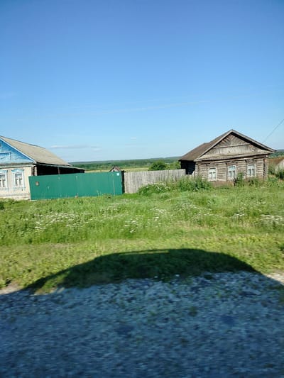 This image depicts a rural Russian landscape under a clear blue sky on a sunny day. Two traditional wooden houses are visible, one with blue trim and a grey roof, and another entirely made of weathered wood with a dark, sloped roof. Both houses have visible windows with white frames. A vibrant green metal fence separates the properties of the houses. Beyond the houses, a wooden fence lines the perimeter of a grassy field dotted with small white wildflowers. In the distance, a treeline marks the edge of the landscape, leading to rolling hills and a hazy horizon. The foreground is dominated by a large, dark shadow cast by an unseen object, likely a vehicle, suggesting the photo was taken while in motion. The ground in the foreground is a blur of grey pebbles and green grass, indicating movement. The scene evokes a peaceful, bucolic atmosphere, characteristic of a summer day in the Russian countryside. There is no visible text in the image.