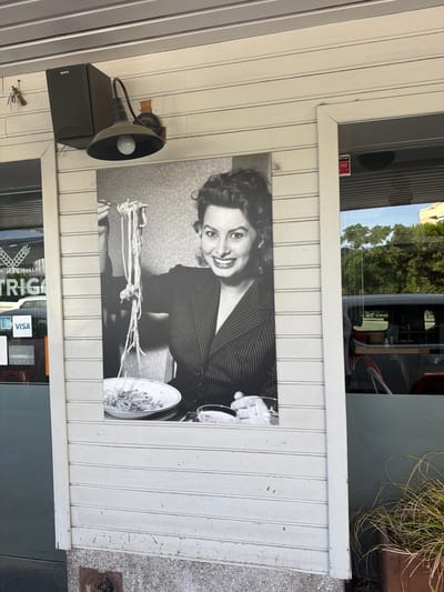This image captures a black and white photograph of actress Sophia Loren on the exterior wall of a building. Loren is depicted holding a forkful of spaghetti, smiling at the camera. The photograph is mounted on a white, horizontally paneled wall. To the left of the photograph, a black speaker with the brand "SONY" and an outdoor light fixture are visible, attached to the wall. Partially visible through a glass storefront to the left of the photograph are the words "TRIGG..." which likely indicates a business name, possibly a bakery given the wheat symbol above it. Payment method decals, including "AM EX" and "VISA", are affixed to the glass. To the right of the photograph, another glass storefront is visible, reflecting the environment outside, including trees and a vehicle. The building's foundation is made of grey aggregate material. The overall scene appears to be a street-level view of a commercial establishment.