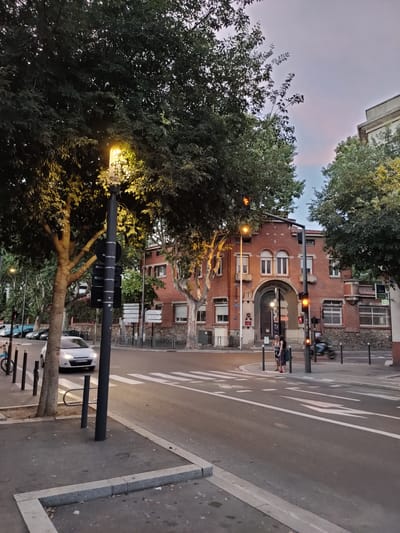 This is a street-level, medium-shot photograph taken in Perpignan, France, featuring a brick building with an arched entrance and an illuminated street lamp in the foreground. The sky is overcast, suggesting dusk or dawn.
The building, a historical structure, is made of red brick with white window frames and decorative stone accents. It appears to be a public building, possibly an administrative or educational institution, with signage visible above the archway. The arched entrance is prominent, with heavy wooden doors.
In the foreground, a black lamppost with a bright yellow light illuminates the scene. The lamppost is positioned on the sidewalk next to a street with a pedestrian crossing. A white car is driving on the street, and two women are standing on the sidewalk near the crosswalk. One woman is wearing a hat and a patterned dress, while the other is in a darker outfit. A motorcycle is visible in motion in the background, suggesting active traffic.
The street is paved and marked with white lines, including a pedestrian crossing. Trees with dense foliage line the street, casting shadows. The sidewalk is made of concrete and stone, with some scattered leaves. The overall atmosphere is urban and relatively quiet, with the artificial light from the street lamp creating a warm glow.
The image captures a moment in a European city, with architecture and street furniture characteristic of France. The specific location within Perpignan is not definitively identifiable from the visual elements alone, but the overall impression is that of a well-maintained urban environment.