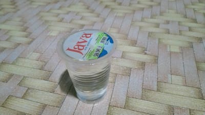 This media file features a close-up, eye-level shot of a clear plastic cup filled with water, resting on a patterned surface that resembles wood planks. The cup is sealed with a white and blue lid that has red lettering. The prominent text on the lid reads "Java AIR MINERAL". Other visible text on the lid includes production details in Indonesian, a barcode, and an SNI logo, indicating it's an Indonesian product. The water level in the cup is approximately half full, with visible ripples on the surface and horizontal indentations on the cup's exterior. The background surface is a mosaic of light brown and cream-colored rectangular shapes arranged in a staggered pattern. The lighting suggests an indoor environment, likely during the daytime, with a soft, diffused light source. There are no people or animals visible. The overall impression is a simple, still-life composition focusing on a single beverage container in a domestic or retail setting. The location context of Kedungpilang, Indonesia, is provided as additional information.