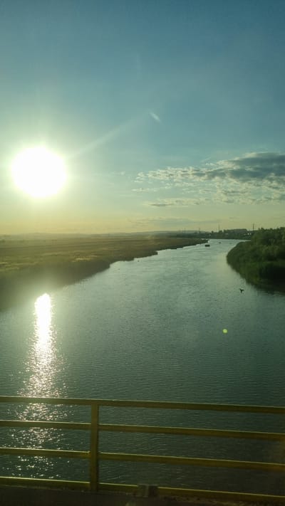 The image captures a wide shot of a river flowing through a rural landscape at sunset, as seen from a bridge railing. The sun, a bright, radiant orb, is positioned in the upper left quadrant of the sky, casting a strong glare and reflecting shimmering patterns on the water's surface. The sky is a gradient of light blue, transitioning to a warmer yellow hue near the horizon, with scattered, wispy clouds.

The river dominates the midground, its surface rippled and reflecting the ambient light. On the left bank of the river, there's a dense area of tall, golden-brown grass or reeds, suggesting a marshy or agricultural area. On the right bank, a line of lush green trees creates a distinct border. In the distance, the landscape appears flat with hints of distant vegetation and possibly some man-made structures like power lines. A few small, dark shapes, possibly birds or boats, are visible on the river.

In the foreground, a yellow metal railing of a bridge is prominently displayed, with its horizontal bars and vertical supports defining the lower edge of the frame. The railing is illuminated by the fading sunlight, adding a touch of color to the foreground. The overall impression is one of serene natural beauty at the end of the day. The provided location of Horia, Romania, suggests this scene is set in a Romanian rural environment.