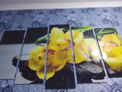 This is a photograph of a five-panel wall art piece featuring a close-up of yellow orchids resting on dark, wet stones. The artwork is displayed on a wall with blue floral wallpaper and a white crown molding at the top. The lighting in the room causes reflections on the surface of the artwork, particularly on the wet stones. The photograph appears to be taken in an indoor setting, possibly a home or a hotel room, located in Namangan City, Uzbekistan, as indicated by the metadata. No people are visible in the image, and there are no discernable actions or interactions taking place.