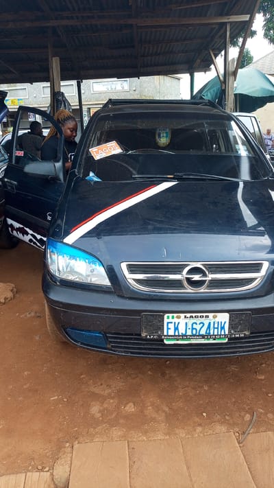 A dark blue Opel car is parked outdoors under a makeshift shelter made of corrugated metal and wood. The car's driver-side door is open, and a woman with blonde braided hair is standing in the doorway, looking down. She is wearing a black t-shirt. A man with a striped shirt is visible in the car's backseat. A license plate on the front of the car reads "FKJ-624HK" and includes "LAGOS" and "A.P.C. Autoservice in Potsdam". Two stickers are visible on the windshield, one orange with "LAGOS" written on it, and another green and gold emblem. The car has a decorative red and white stripe on its hood, and the headlights have a blue tint. The ground is reddish-brown dirt with scattered stones and some paving stones. In the background, a building with large windows is visible, along with some greenery and trees. The overall scene appears to be a repair shop or a lot where vehicles are parked. The specific location in Kafanchan A, Nigeria, is not explicitly indicated in the image.