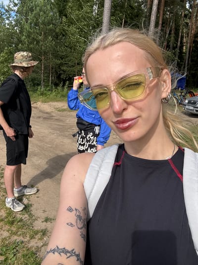 This is a close-up, rotated selfie of a young woman with blonde hair, wearing large yellow-tinted sunglasses. She has silver hoop earrings in her left ear and a small stud. Her lips are painted with a reddish-pink lipstick. She is wearing a black top with white and red trim. Behind her, partially visible, are two other individuals. One person is wearing a blue jacket and a black t-shirt with white text. The other person is wearing a camouflage hat and a black shirt. They appear to be outdoors in a wooded area with dirt ground and grass. The background shows trees and what looks like a parked car and bicycles, suggesting an outdoor recreational setting. The lighting indicates it is daytime with bright sunlight, creating shadows. The scene appears casual and relaxed. The text on the black t-shirt reads "Ego". There is also some text on the license plate of the car which appears to be "M990 KT 67". The location is contextually identified as Desnogorsk, Russia.