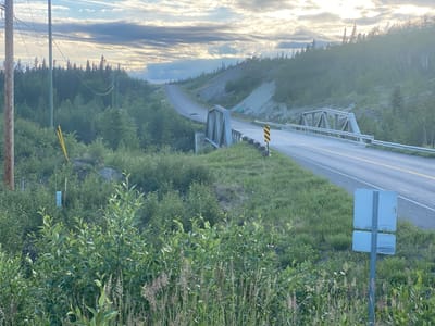 In Haines Junction, Canada, a tranquil scene unfolds under a cloudy sky. The image captures a two-lane asphalt road winding its way through a lush green landscape. The road is bordered by guardrails and shoulders of grass and brush. On the left side of the road, a rustic wooden utility pole stands tall, adorned with wires that stretch towards the horizon. Further down, a small metal truss bridge crosses a ravine, adding a touch of industrial charm to the natural setting.
The foreground is dominated by a vibrant array of greenery, with bushes and tall grasses swaying gently. In the distance, the road ascends a hill, disappearing into the misty expanse. The surrounding terrain is characterized by gentle slopes covered in dense forestation, with a scattering of deciduous trees and evergreen conifers. The sky above is a dramatic canvas of swirling clouds, with patches of sunlight breaking through, casting a soft glow on the landscape. A white sign with a rectangular shape and two smaller stacked rectangles is affixed to a metal pole on the right side of the road. A yellow marker with a diagonal line is visible in the brush on the left.
