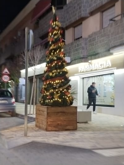 A Christmas tree stands in a wooden planter outside a store. The tree is decorated with lights and ribbon. A man in a black hoodie and dark pants walks in front of the store. The store has a bright sign that says "FARMACIA" in illuminated letters. A gray car is parked on the side of the road. A street sign is visible. The scene is at night.
