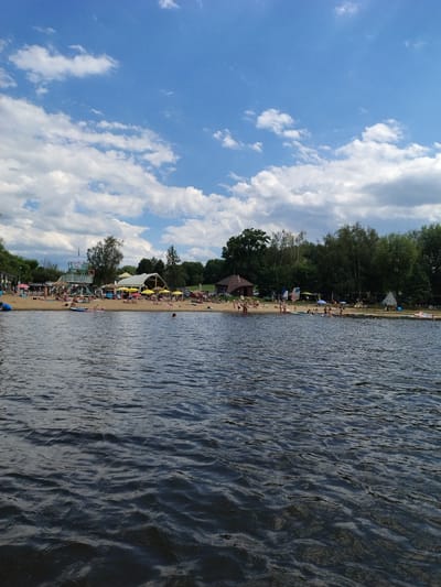 This is a wide-angle, eye-level shot of a busy lake beach on a cloudy but bright summer day, located at the Radslavski rybnik (Radslav Pond) in Radslav, Czechia. The foreground is dominated by dark, rippling lake water. The midground features a sandy beach populated by numerous sunbathers and swimmers. Several yellow and white umbrellas provide shade. Notable structures on the beach include a multi-story lifeguard tower with a light blue and white structure, a large white tent or canopy structure, and a small, reddish-brown building with a dark roof towards the right. Behind the beach, dense green trees form a treeline. In the sky, scattered white cumulus clouds are visible against a bright blue backdrop. The scene depicts a recreational area with active human engagement.