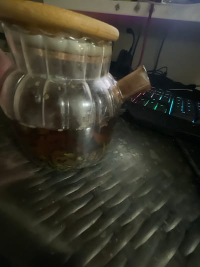 A close-up, dimly lit image shows a glass teapot with a wooden lid and a spout made of wood or bamboo. The teapot is filled with a dark liquid, likely tea, and appears to contain some tea leaves at the bottom. It sits on a dark, textured surface, possibly a desk, which has a ribbed pattern. In the background, a computer keyboard with illuminated keys is visible, suggesting an indoor setting. The overall impression is one of a cozy, perhaps late-night, moment of enjoying tea while working on a computer. The location is identified as Bol'shoe Zaborod'e, Russia.