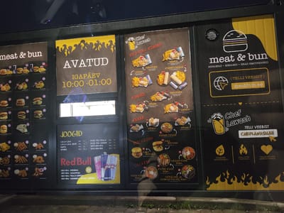 The image shows a nighttime exterior view of a fast-food restaurant called "meat & bun" and "Chef Lawash" located in Laagri alevik, Estonia. The menu boards are illuminated, and the restaurant appears to be a drive-thru or walk-up window. The menu boards display various food items, including burgers, kebabs, shawarmas, bowls, and drinks, with prices listed in Euros. The "meat & bun" menu features a logo of a hamburger and lists categories like "BURGERID, KEBABID, HEAD EMOTSIOONID". The "Chef Lawash" menu has a logo of a wrapped food item and lists categories like "RED BULL KOMBO", "DÖNER", "SHAURMA", "KEBAB", "BURGERID", and "SALATID". There are also Red Bull energy drink cans displayed prominently on the menu. The overall scene is dark, with some ambient light reflecting off the glass, suggesting it might be raining or there is condensation on the window. No people are visible in the frame.