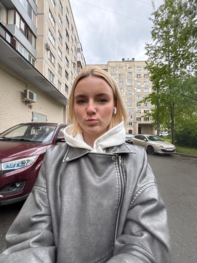 A young woman with blonde hair is taking a selfie outdoors. She is wearing a white hoodie and a silver jacket. In the background are apartment buildings, trees, and parked cars. One of the cars is red, another is silver. The license plate number visible on the silver car is O 977 TT 47. The building facades appear to be in Novoe Devyatkino, Russia.