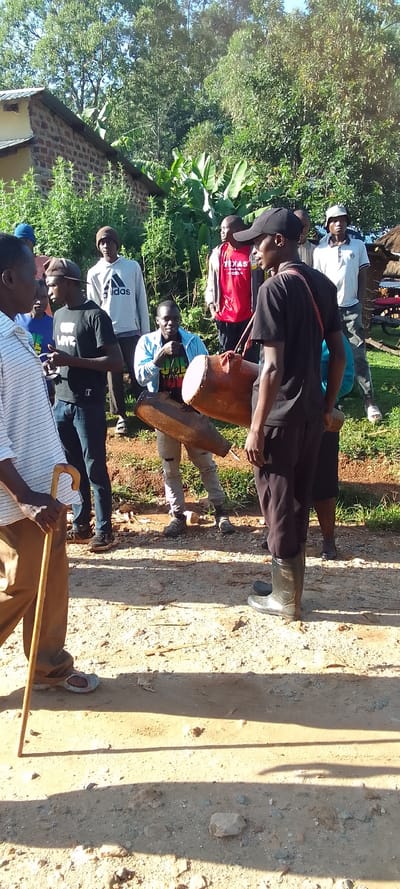 The image shows a group of people standing on a dirt road in Isukha ICHINA, Kenya. The coordinates (latitude: 0.352, longitude: 34.782) place the scene in a rural area. In the foreground, a man is walking, holding a wooden walking stick. He is wearing a white, striped shirt and brown pants. He is wearing sandals. In the middle of the image are six men. One man is holding a wooden drum. He is wearing a black T-shirt and black pants. The other man is holding a large, brown drum. He is wearing a black shirt, black pants, and black boots. The others are standing around them, watching. They are wearing casual clothes, with three of them wearing hats. Behind the group is a brick building with a green roof. There is a small, brick building with a green roof. The environment is rural and dusty, with trees and foliage in the background. The scene appears to be an informal gathering of people, possibly a community event.