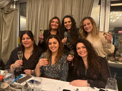 Six smiling women pose with champagne glasses indoors. The woman in the center back wears a tiara and a "32 and Fabulous" sash. They are gathered around a table set with wine glasses, cutlery, and confetti. Neutral-colored curtains hang in the background. One woman in the back has her arm around the woman in the front.