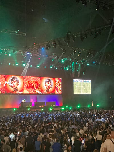 This image captures a concert or large event at the Stade Pierre-Mauroy in Villeneuve-d'Ascq, France. A large crowd of people is gathered in the foreground, facing a stage area. Above the crowd, large screens display visuals, with one screen featuring stylized portraits of a woman and the number "1970" in a prominent, colorful font. Another smaller screen displays a soccer match in progress. The stage itself is illuminated by a variety of colored lights, with green lights being particularly prominent in the upper portion of the image. Lighting rigs and speaker systems are visible suspended above the stage. The overall atmosphere appears to be energetic, with the crowd suggesting a significant attendance.