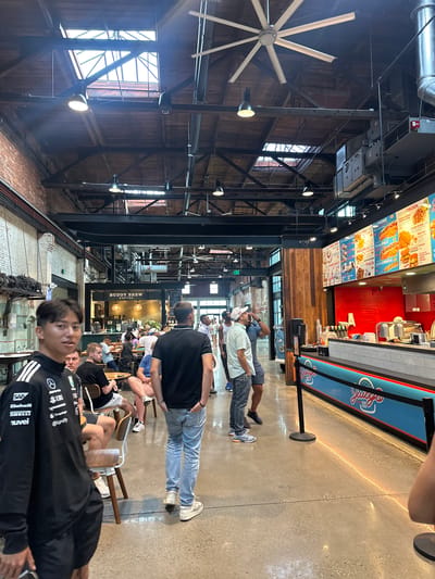 The image captures an interior scene, probably in a spacious industrial-chic dining establishment in Tampa, United States. Patrons stand in line at "Spole's", a casual food establishment which prominently features menu items like chicken, mozzarella fries, and shakes. A group of people are seated at tables in the dining area further inside the building at "Buddy Brew Coffee". The architecture features high ceilings with exposed beams and industrial-style lighting fixtures. A large fan is visible near the ceiling. A man wearing a black sweatshirt with corporate logos faces the camera. People are standing, seated, and waiting in line. The atmosphere appears to be casual and social.