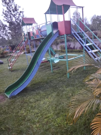 A colorful playground structure dominates the scene. It features a blue and green slide curving towards the left, along with a cylindrical component connecting two elevated platforms. One platform is accessible by red stairs, the other by blue stairs. All metal supports are painted green. The structure stands on a green lawn, with a palm tree on the right and trees in the background under an overcast sky.