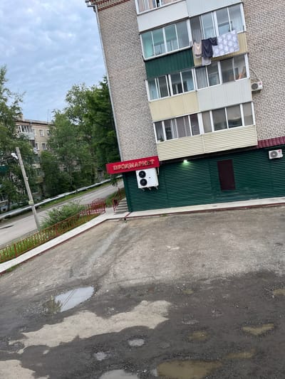 The image shows a brick apartment building with a green storefront on the ground floor. The storefront has a red sign that reads "ПРОДМАРКЕТ," which translates to "Prodmarket" or "Food Market." There are two air conditioning units mounted on the exterior wall of the building, one above the storefront and another on the side. A dark-colored door is visible on the green storefront. In front of the building, there is a paved area with several potholes filled with water. A red metal railing lines a sidewalk adjacent to the paved area, and beyond that, there is a road with a large metal pipe running alongside it. Trees and another apartment building are visible in the background. The sky is overcast. The scene is likely an urban or suburban environment in Magdagachi, Russia.
