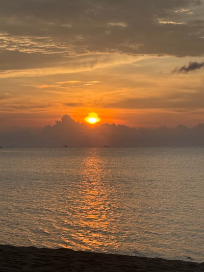 The image captures a sunset over the ocean in Baan Wang Ta Kien, Thailand. The sun is positioned near the center, partially obscured by clouds, creating a warm, orange glow across the sky and reflecting on the water's surface. The sea exhibits rippled texture with light reflecting on the surface. A coastline is visible to the right of the body of water. Small boats dot the horizon. The scene is bathed in a golden light, indicative of the time of day.