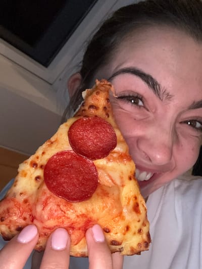 A close-up image depicts a female holding a triangular slice of pepperoni pizza in front of her face. The woman is positioned on the right side of the frame, partially obscured by the pizza. She has dark hair, pronounced eyebrows, and is captured mid-facial expression, characterized by a wide smile or grimace, revealing her teeth and pronounced crow's feet wrinkles around her right eye. Her left hand, with light pink manicured fingernails, supports the pizza from below the crust. The pizza slice features a golden-brown crust, melted cheese, and two distinct circular pepperoni slices. The background is dark and out of focus, showing a white window frame at the upper left, indicating an indoor setting in low light conditions within the city of Villeurbanne, France.