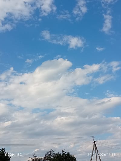 The image depicts a wide-angle view of a cloudy sky with a clear blue expanse. Cumulus clouds, varying in density, are scattered across the frame, suggesting a partly cloudy day. In the lower portion of the image, a silhouetted treeline is visible, hinting at a rural or natural setting. Power lines stretch horizontally across the middle ground, with a utility pole featuring insulators in the right half of the frame. The scene appears to be outdoors during daylight hours, likely late afternoon given the lighting.
Visible text includes "REDMI 15C" in the bottom left corner and a date and time stamp "24.06.2026 17:58" in the bottom right corner. The location context provided is Mykolaivka, Ukraine. No people or specific activities are discernible. The overall mood is calm and serene, characteristic of a pleasant day with a dynamic sky.