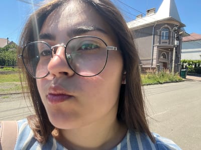 The media file is a close-up, upside-down selfie of a young person's face, taken outdoors on a sunny day. The subject is wearing round, metal-rimmed glasses and a blue and white striped top. Their brown hair is visible framing their face. The background shows a street and a residential area with houses and fences, suggesting a suburban or semi-rural setting in Chahor, Ukraine. The lighting indicates it is daytime, likely late morning or afternoon, given the bright sun. There is no discernible text in the image. The focus is primarily on the subject's face and the immediate surroundings, with the tilted perspective adding a dynamic element to the composition. The overall mood appears casual and relaxed.