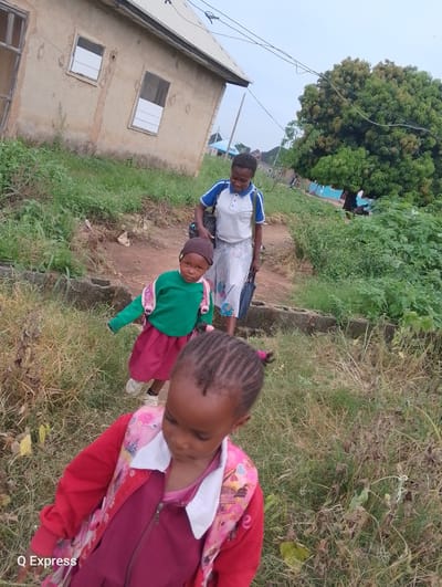In Sabojibi, Nigeria, three individuals are present near a simple, weathered building with two windows. The building appears to be constructed of concrete and has a corrugated metal roof. Surrounding the structure is overgrown vegetation and dry grass, suggesting an unkempt or rural setting.

In the foreground, a young girl is captured from behind, facing away from the camera. She wears a burgundy dress and a bright red jacket adorned with a pink floral pattern.

Behind her, another young girl, looking directly at the camera, is dressed in a green long-sleeved shirt and a burgundy skirt. She has a dark beanie on her head and a pink backpack slung over her shoulders.

Further back, a young woman walks away from the camera, looking over her shoulder and smiling. She is wearing a white polo shirt with blue trim and a patterned skirt. She carries a dark bag in her right hand. The scene suggests the group may be walking or preparing to walk somewhere, possibly to or from school, given the presence of backpacks. The overall environment is modest and suggests a developing area.