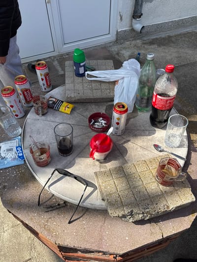 An outdoor scene in Sotirya, Bulgaria, depicts a collection of discarded and in-use items on and around a stone or concrete table, illuminated by direct sunlight.
**Location:** The setting is an outdoor patio or courtyard against a light-colored building wall. A white door or window frame with a handle is visible in the background, along with a grey downspout affixed to the wall. The ground consists of light grey paving stones.
**Objects and Arrangement:** The central feature is a roughly circular, rustic stone or concrete table with a textured surface. On the table, various items are scattered, suggesting recent or ongoing consumption:
* **Beverages:** Four partially consumed or empty 0.5-liter Stella Artois beer cans, some with a red heart design, are present. A 1.5-liter plastic Coca-Cola bottle, partially full, stands near an empty clear green glass bottle.
* **Drinking Vessels:** Two clear glass mugs with "CAPPUCCINO" branding; one contains a spoon and brown residue, the other is mostly empty. Two additional clear glass tumblers are visible, one empty, one containing a dark liquid. A dark, opaque glass or plastic cup also contains a dark liquid.
* **Smoking Paraphernalia:** A red circular "LEADER" branded ashtray is filled with numerous cigarette butts and ash. A yellow and black cigarette pack, displaying a Bulgarian health warning, lies open next to a clear plastic bag containing OCB brand cigarette filters.
* **Other Items:** A red-topped, white-based sugar dispenser with a small spoon is on the table. A pair of black sunglasses, folded, rests near the table's edge. A crumpled white plastic bag and a green aerosol can are positioned on a separate concrete block next to the table. A loose silver spoon is also present.
**People:** The lower torso and legs of an individual wearing grey sweatpants and a dark jacket are visible in the upper left of the frame, indicating human presence near the table. No direct actions or interactions are depicted for this individual.
**Environment:** The ground around the table is paved with large, light-colored concrete squares. The strong sunlight casts sharp shadows from the objects, indicating a clear, bright day.
