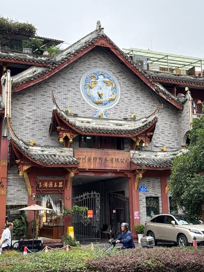 The image captures a street scene featuring a multi-story building with traditional Chinese architectural elements on Citang Street (祠堂街) at address 157 in Qingyang District, Chengdu, China. The building's facade is constructed of light grey bricks, adorned with red wooden structures, tiled roofs with upturned eaves, and gold decorative accents. A large circular blue and gold mosaic depicting two dragons is prominently centered on the upper facade.

Below the dragon mosaic, an arched entrance frames a metal gate, with a weathered brown sign above it displaying Chinese characters reading "四川国防印务有限公司" (Sichuan National Defense Printing Co., Ltd.). To the left of the archway, a ground-floor shop entrance is visible, featuring a dark red facade with gold trim. A sign above this shop reads "玉满源玉翠" (Yuman Yuan Yucui). An open umbrella shades a small outdoor display or seating area near this shop, with potted plants surrounding it.

On the street level, a short, trimmed hedge or planter separates the sidewalk from the building. To the far left, a man in a light-colored shirt is partially visible, standing near a cart with wheels and a red traffic cone. To the right of the building's main archway, a blue street sign clearly shows "祠堂街 157号" (Citang Street No. 157). A red "禁止停车" (No Parking) sign is attached to the metal gate of the main archway. Further right, an elderly person with grey hair, wearing a blue jacket, is seated on and operating a three-wheeled mobility scooter, which has a silver storage container on its rear. Behind the scooter, a gold-colored Lexus RX SUV is parked facing the viewer. The upper sections of the building's roof show various structures, including what appears to be a light green corrugated metal canopy and utility equipment or water tanks, amidst some overgrown greenery. The sky above is overcast and light grey.