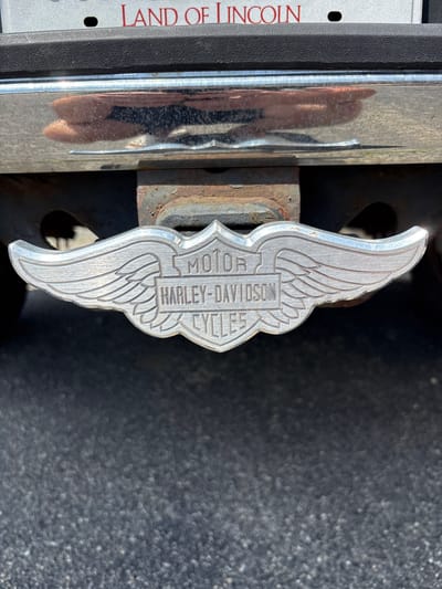A close-up, eye-level shot shows a silver Harley-Davidson Cycles logo attached to a hitch receiver on the back of a vehicle. The logo is wing-shaped and features the words "MOTOR", "HARLEY-DAVIDSON", and "CYCLES" within a shield design. The hitch receiver is mounted to a dark-colored frame underneath the vehicle's bumper, which is chrome and reflects a blurry image of what appears to be hands and fabric. Above the bumper, a portion of a license plate is visible with the words "LAND OF LINCOLN" in red lettering. The background consists of a dark asphalt surface. The scene is outdoors on a bright day.
