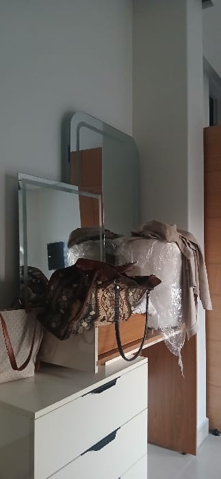 The image displays an interior room, presumed to be in Ashrafia, Syria, containing various furniture and personal items. In the foreground on the left, a white dresser with at least three visible drawers featuring recessed dark handles is present. On top of this dresser, a light-colored bag with brown straps and a subtle pattern is partially visible. Centrally, a wooden vanity or desk supports a large bag with an intricate dark and light pattern, accompanied by dark straps. Behind this patterned bag, a rectangular mirror with a thin frame is positioned vertically. To the right of the rectangular mirror, another mirror with a rounded top edge is affixed or leaning against a light-colored wall. A large object, potentially furniture, is covered in crinkled clear plastic sheeting, over which a light-brown or beige garment is casually draped. The background consists of light-colored walls, a prominent vertical white column on the right, and a light-brown wooden door or panel situated further right. Reflections in the mirrors show portions of the room, including a dark rectangular object. No individuals are present in the scene.