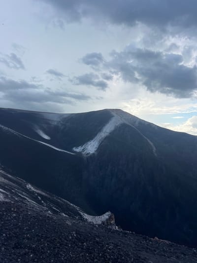 The image depicts a steep, dark, volcanic landscape under a cloudy sky. The primary subject is a mountain, likely a volcano, with a dark, rocky surface. A prominent feature is a lighter-colored streak running down the side of the mountain, possibly indicating a path or a geological formation. The foreground is also composed of dark, loose volcanic rock. In the distance, the sky is filled with dramatic, heavy clouds, with patches of brighter sky and sunlight breaking through. The overall impression is one of a rugged and imposing natural environment. The scene appears to be set during the daytime, but the cloudy conditions suggest it could be overcast or approaching stormy weather. The specific location, Rota, Nicaragua, is known for its volcanic activity and dramatic landscapes, which aligns with the visual cues in the image. There is no visible text in the image.