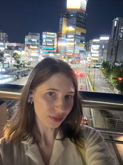 This is a nighttime selfie taken by a woman with light brown hair who is wearing a light-colored shirt and a light purple earbud. She is standing on a pedestrian overpass. In the background, there is a cityscape with numerous buildings adorned with bright neon signs, indicating a commercial area. A street with traffic, including cars and motorcycles, is visible below the overpass. The overall scene depicts a vibrant urban environment at night. The image is tagged with "Uijeongbu-si, South Korea".
