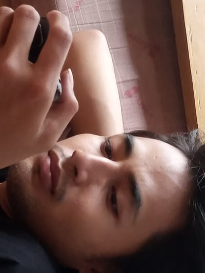 A male individual is depicted lying on his back, with his head tilted to the right, in an indoor setting within the city of Muka, Indonesia. His dark hair is visible, and he possesses dark eyes and light facial stubble. He is wearing a dark-colored garment, likely a shirt. His left hand is positioned in the upper left quadrant, holding a dark, rectangular electronic device, possibly a smartphone or remote control. A bare human leg is visible to the right of his hand, resting on a patterned surface. The background consists of a light-colored, possibly faded pink, fabric surface with faint, repetitive motifs, consistent with bedding or a cushion. A vertical wooden structure, likely furniture, is present along the right edge of the frame. The individual's gaze is directed towards the camera.