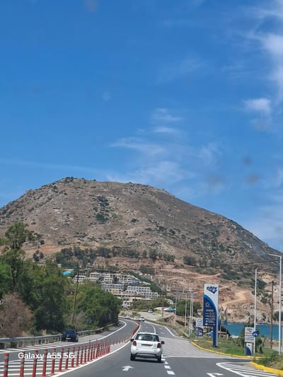 The image depicts a coastal road curving along the base of a steep, rocky hillside in Gazi Municipal Unit, Greece. A white car is visible driving on the road, while a dark vehicle is parked near a guardrail painted with white and red stripes. A gas station sign is positioned at the roadside. The hillside is partially vegetated, with buildings clustered towards the lower slopes. The sky is blue with scattered clouds.