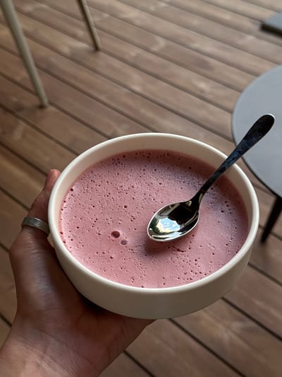 A hand, adorned with a silver ring, holds a white bowl filled with a frothy, pink substance, possibly a mousse or a smoothie. A silver spoon rests in the bowl, reflecting the surrounding environment, which appears to be an outdoor setting with wooden planks forming the surface. The scene is lit by natural light, suggesting it's daytime. The background is blurred, but hints of outdoor furniture, specifically a grey circular table and possibly some railings, can be discerned. The wooden planks are oriented diagonally, creating a dynamic background pattern. The overall mood is casual and relaxed, capturing a moment of enjoying a refreshing treat. No text is visible in the image.