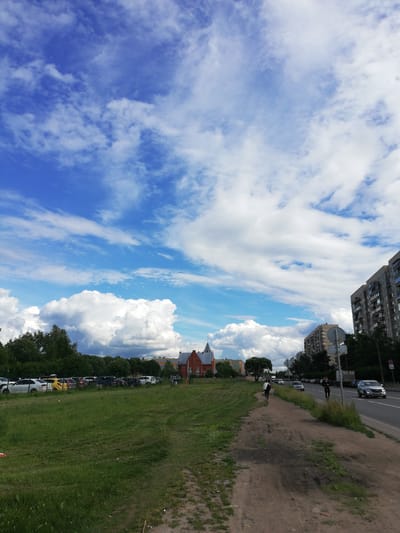 The image depicts a street scene in Kolpino, Russia, under a partly cloudy sky. In the foreground, a dirt path runs alongside a grassy field. Beyond the field, a row of parked cars is visible, some white, one yellow, and others darker shades. Further in the background, several buildings can be seen, including a red brick building with a pointed roof, and a tall, multi-story apartment building on the right side of the frame. Trees line the edge of the field and the area behind the parked cars.
A paved road runs parallel to the dirt path, with several cars driving on it, including a silver SUV. Two people are walking on the dirt path, one with a backpack. Another person appears to be riding a scooter. The sky is a vibrant blue, dotted with numerous white, fluffy clouds. The sunlight suggests it is daytime, likely afternoon, given the shadows. The overall atmosphere is casual and urban, with a hint of nature in the grassy field and trees. No visible text is present in the image.