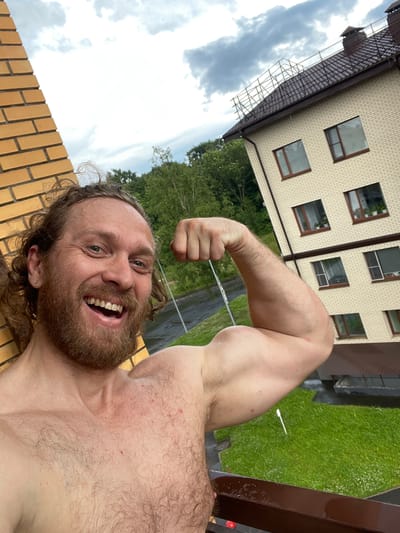 In this selfie, a man with long, curly brown hair and a beard poses on a balcony, flexing his bicep. He is shirtless, revealing a hairy chest and shoulder. The man is smiling broadly, with his mouth open. In the background, a multi-story brick building with dark brown trim and windows can be seen. Trees and a cloudy sky complete the outdoor setting. The man appears to be in the settlement of Kornevo, Russia.