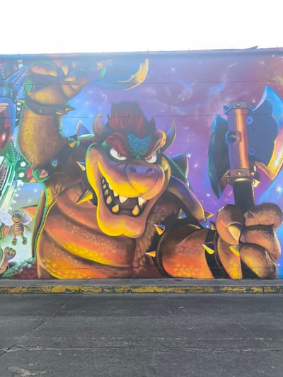 A mural featuring Bowser from the Super Mario franchise is painted on a wall in the city of La Victoria, Peru. The mural depicts Bowser holding a large axe with a starry background. A yellow curb runs along the bottom of the wall, with a paved street visible in the foreground.