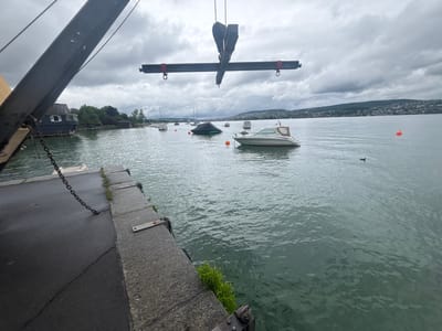 The image depicts a waterfront scene in Kilchberg (ZH), Switzerland, under a cloudy sky. A crane, partially visible in the upper left and center, suspends a horizontal beam with hooks. In the water, several boats are visible, including a white motorboat in the center, some covered boats, and other smaller vessels in the distance. Orange buoys are scattered in the water. A dock extends into the water in the foreground. The background shows a distant shoreline with buildings and trees. A duck floats in the water near the center-right.