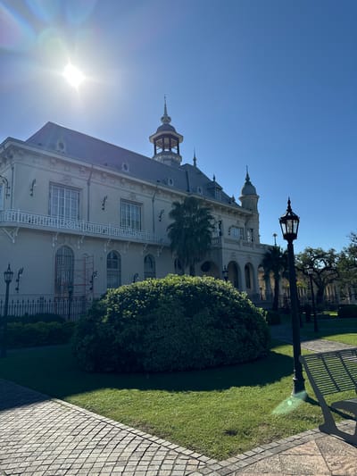 The image is an exterior shot of the Tigre Art Museum, located in the city of Tigre, Argentina. The museum is a large, ornate building with a light-colored facade, a dark blue roof, and several turrets. In front of the museum is a green lawn with a large bush and a black metal lamppost. The sky is blue, and the sun is shining brightly.