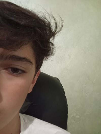A close-up image shows the left side of a person's face and head. The person has dark brown hair styled with some strands sticking up, a defined eyebrow, and a brown eye. The visible part of the face shows fair skin. The person is wearing a white shirt. A dark, possibly black, chair or object is partially visible behind the person. The background is a light-colored, textured wall. The location in the city of Malko Chochoveni, Bulgaria cannot be determined from the image.