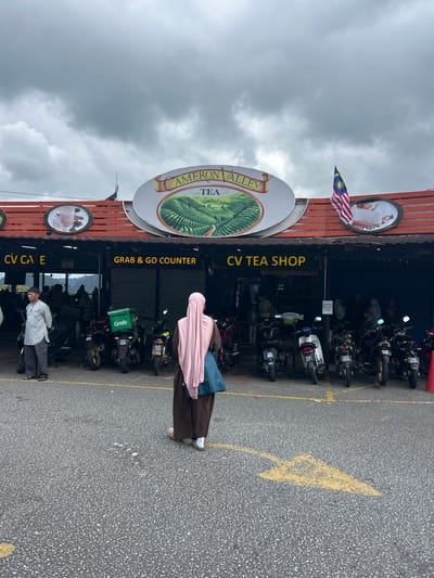 The image shows an outdoor scene in front of a building labeled "CAMERON VALLEY TEA SHOP". The building has an orange roof and a sign depicting tea plantations. There are various shops or counters visible, including "CV CAFE" and "GRAB & GO COUNTER". A Malaysian flag is displayed on the roof. In the foreground, a person wearing a pink hijab and brown attire walks away from the camera. Several motorcycles are parked in front of the building. The ground is asphalt, with yellow painted markings. The sky is overcast and cloudy. The location is identified as Tanah Rata, Malaysia, known for its tea plantations.